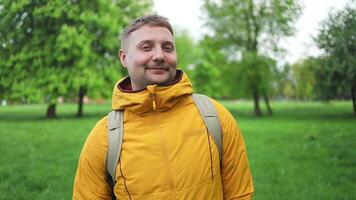 Handsome Caucasian tourist man outdoors in a yellow sports jacket and backpack, looking at the camera. Male relaxes and enjoys the fresh air in a city park. Man smiling, looking in the camera video