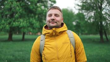 Handsome Caucasian tourist man outdoors in a yellow sports jacket and backpack, looking at the camera. Male relaxes and enjoys the fresh air in a city park. Man smiling, looking in the camera video