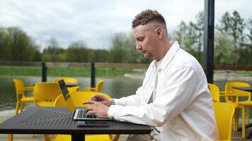 lado ver de caucásico hombre quien se sienta en ciudad al aire libre café en tiempo de día y utilizando el ordenador portátil para el remoto trabajar. persona de libre dedicación empresario mecanografía texto en el teclado en cafetería video