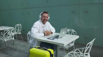 Friendly Caucasian 30s man sits in airport terminal using laptop talking via call. Male waiting for flight, browse internet, social media, online shopping. video