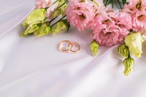 Two gold classic wedding rings on a white satin background with fresh pink austoma flowers. A copy space. postcard. invitation. cover. photo