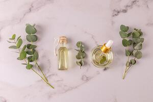 Bottle and bowl with pipette of natural organic eucalyptus oil on a white marble background with eucalyptus branches. Upper view. Spa treatment. photo