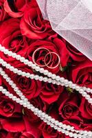 Background image for wedding design or mockup. Two gold wedding rings on a red roses bouquet background. white bridal veil. Pearl Beads. Top view. photo