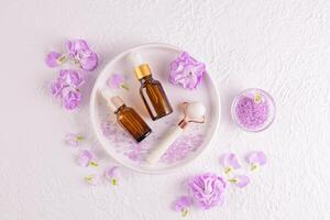 Two Dropper Bottles With Natural Skin Care Product, Oil For Massage, Stone Roller Massager On White Round Tray Among Flowers. Top view photo