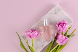 An elegant bottle of eau de parfum or women's perfume lies on a marble rectangular podium with tulips. Fragrance presentation. Top view. photo