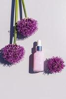 A bottle with a delicate serum for the care of young skin on a light background with lilac decorative Allium flowers. Care and beauty concept.. photo