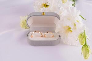 Two gold wedding classic rings in a gray velvet box on a white satin fabric with fresh white flowers. Wedding concept. Front view photo