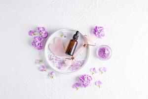 Cosmetic dropper bottle with natural organic product for care and massage on ceramic plate with roller for massage. lilac delicate flowers. photo