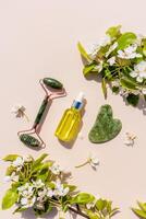 conjunto de natural jade cosmético masajeador y gua sha raspador, petróleo para linfático drenaje masaje en beige vertical antecedentes. ramas de un floración. foto
