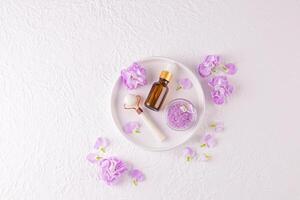 Cosmetic set for spa and skin care, massage. Stone roller, bath salts, massage oil on ceramic white tray. top view. lilac delicate flowers photo