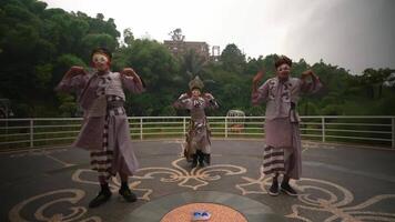 Three performers in traditional Japanese masks and costumes enact a scene outdoors with greenery and a building video