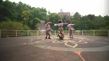 Three dancers in traditional attire performing a synchronized routine on an outdoor circular stage with lush greenery and a historic building video