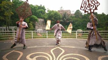 Performers in traditional costumes dancing on an outdoor stage with a lush green backdrop and a castle-like building video