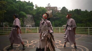 Performers in traditional theatrical costumes and painted masks engaging in a dramatic outdoor performance, with a lush green cliff video