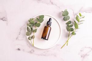 Cosmetic bottle with natural face and body care product with eucalyptus oil extract on a white round tray. Marble background. Blank product mockup. photo