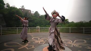 dos intérpretes en tradicional mascaras y disfraces bailando en un al aire libre etapa con verdor y esculturas video