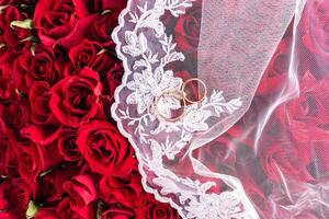 Background image for wedding design or mockup. Two gold wedding rings on a white bridal veil. Top view. red background roses bouquet. A copy space photo