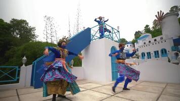 Performers in vibrant traditional costumes dancing in front of a whimsical blue and white building, evoking a festive video