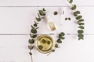 Cosmetic product based on natural eucalyptus oil on a wooden white background with eucalyptus branches and a bowl of organic oil. Top view photo
