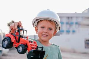 A little boy in a white helmet is holding a toy red loader. A child on the background of a construction site plays with a typewriter and smiles. The profession is a builder. photo