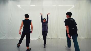 Three young adults practicing dance moves in a studio with white walls, one in focus at the front with two video