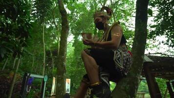 Man in traditional Javanese costume and mask sitting in a lush forest, reading a book, showcasing cultural heritage video