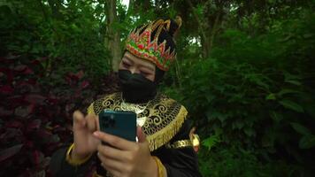 Person in traditional ethnic attire and mask using a smart phone in a lush green forest setting video