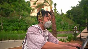 Side view of a focused woman wearing a mask and using a laptop outdoors with lush greenery and a historical building in the background video