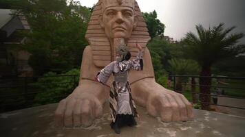 Tourist posing for a photo in front of a large stone sculpture of a head with crossed arms in a lush, green park setting. video