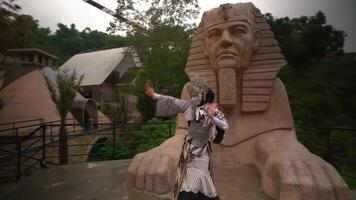 A person in a patterned outfit stands in front of a large sphinx statue with lush greenery in the background, creating a unique juxtaposition video