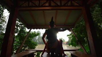 Silhouette of a person practicing yoga in a serene pavilion overlooking a tranquil lake, surrounded by lush greenery video