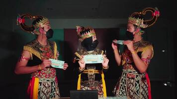 Tres individuos en tradicional balinés atuendo y protector cara mascaras utilizando moderno tecnología, mezcla cultural patrimonio video