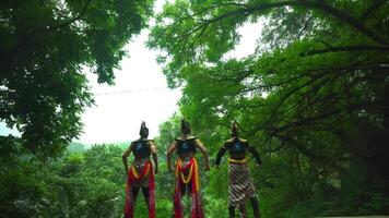 Three performers in traditional costumes and headgear stand amidst lush greenery, their backs to the camera, evoking a sense of cultural performance video