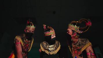 Three performers in traditional Javanese dance costumes and masks, with intricate headgear, captured in a dimly lit setting video