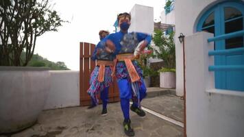 twee blij dansers in kleurrijk outfits en gezicht maskers het uitvoeren van een traditioneel dans Aan een straat video