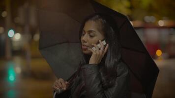 jovem mulher falando em Móvel telefone lado de fora dentro a chuva às noite video