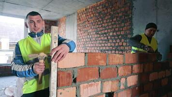 bouw arbeider gebouw steen muur, een bouw arbeider vervelend een rood moeilijk hoed en geel veiligheid hesje is gebouw een steen muur Aan een bouw plaats video