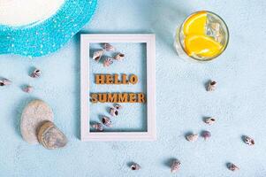 Hello summer text in photo frame, cocktail, hat, seashells and stones on blue top view