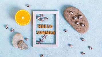 Hello summer text in photo frame, orange, seashells and stones on blue top view web banner