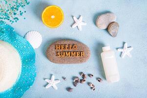 Hello summer text on stone, hat, suntan lotion, orange, starfish and seashells on blue background top view photo