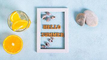 Hello summer text and seashells in photo frame, cocktail and orange on blue top view web banner