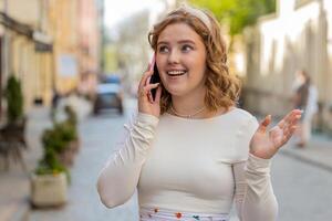 Happy young woman having remote conversation talking on smartphone good news gossip in city street photo