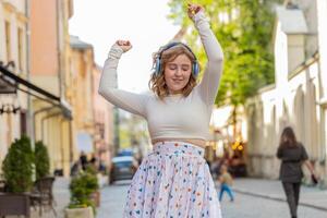 Happy smiling redhead woman in wireless headphones listening music dancing outdoors in city street photo