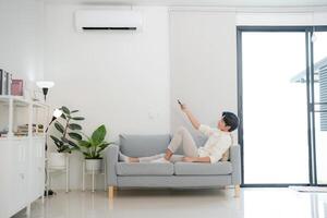 joven hombre disfrutando comodidad utilizando remoto controlar para aire acondicionamiento a hogar foto