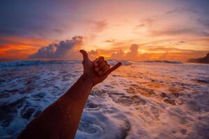 Hand with shaka symbol and waves with sunset in ocean. Lifestyle surfing concept photo