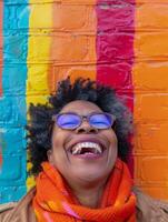 A woman in electric blue eyewear laughs joyfully by a colorful brick wall photo