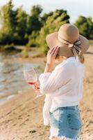 A girl in a straw hat with a glass of wine sits on the beach at sunset. photo