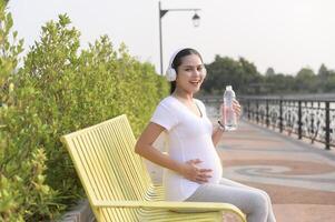 hermosa embarazada mujer Bebiendo botella de puro agua en el parque, sano y activo el embarazo estilo de vida concepto. foto