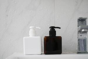 Two square-shaped bottles, one white and one brown, with black pumps, sit on a wet sink in front of a white-tiled wall. photo