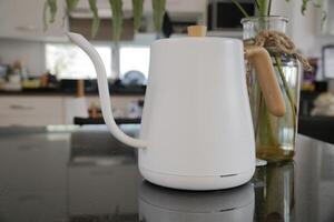 A white kettle with a wooden handle sits on a black counter next to a clear vase with a plant in it. photo